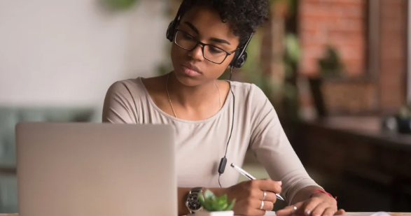 Woman with headphones watching webinar on laptop and taking notes